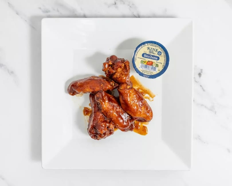 Four saucy chicken wings on a white plate with a side of Ken's Bleu Cheese dressing.