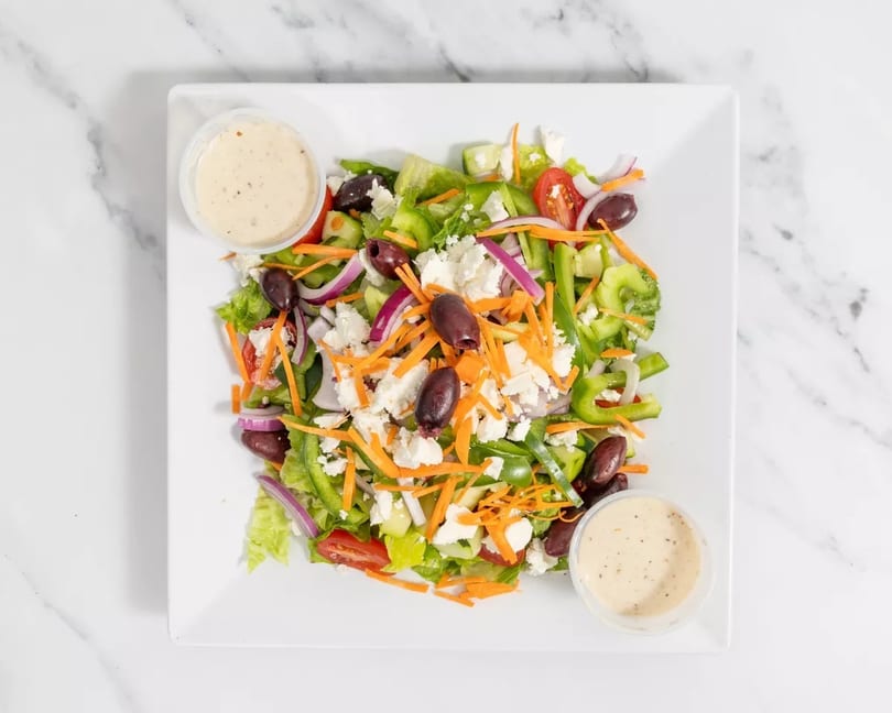 A fresh Greek salad with lettuce, tomatoes, olives, feta, and dressing on a white plate.