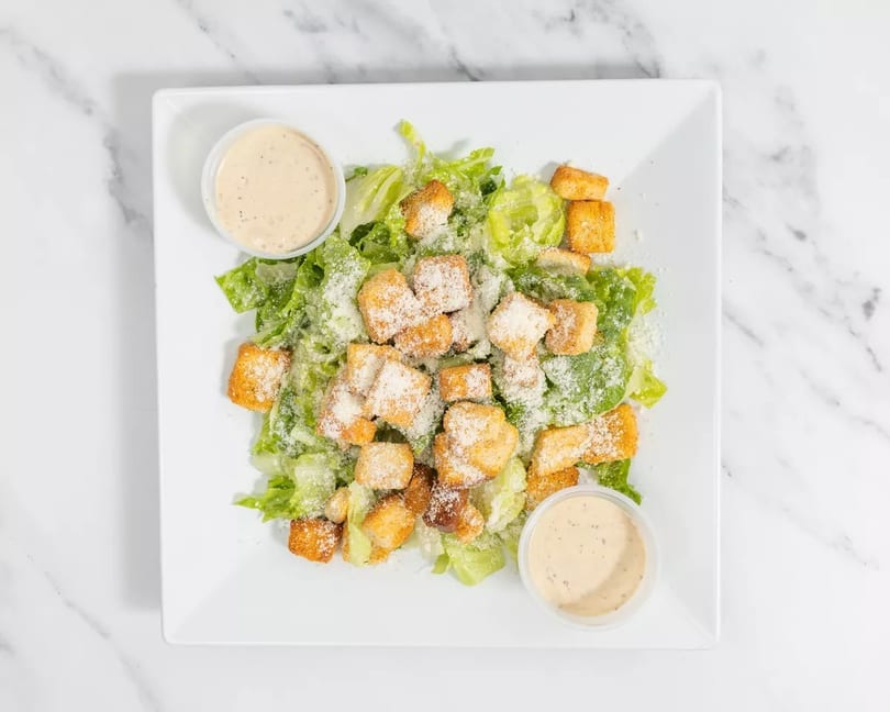 Caesar salad with romaine lettuce, croutons, parmesan cheese, and two cups of dressing on a white plate.