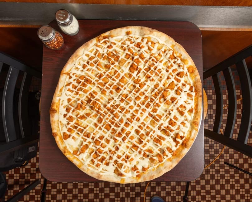 Overhead view of a large buffalo chicken pizza with white sauce drizzle on a dark wooden table.