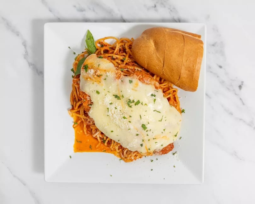 Chicken parmesan with melted cheese, spaghetti, and a side of bread on a white plate.